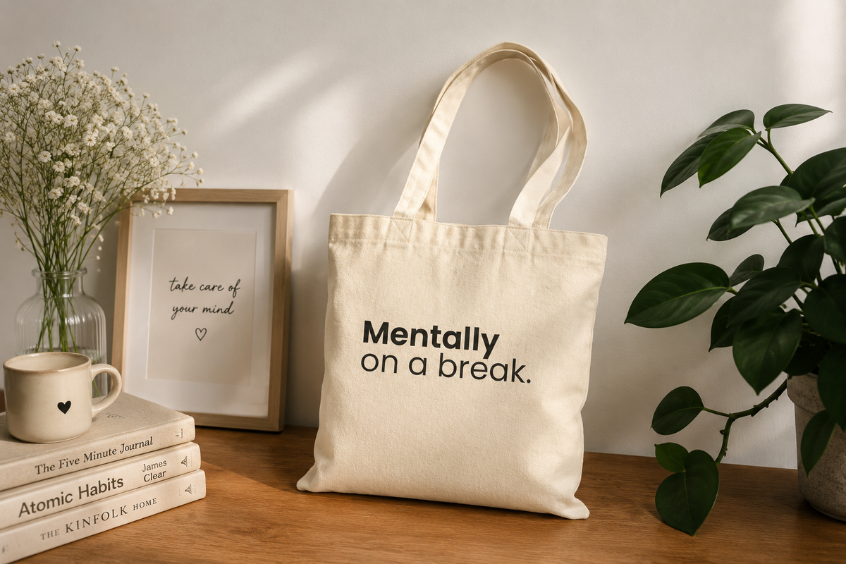 Beige canvas tote bag with "Mentally On a Break" statement design styled with books, coffee, and a houseplant
